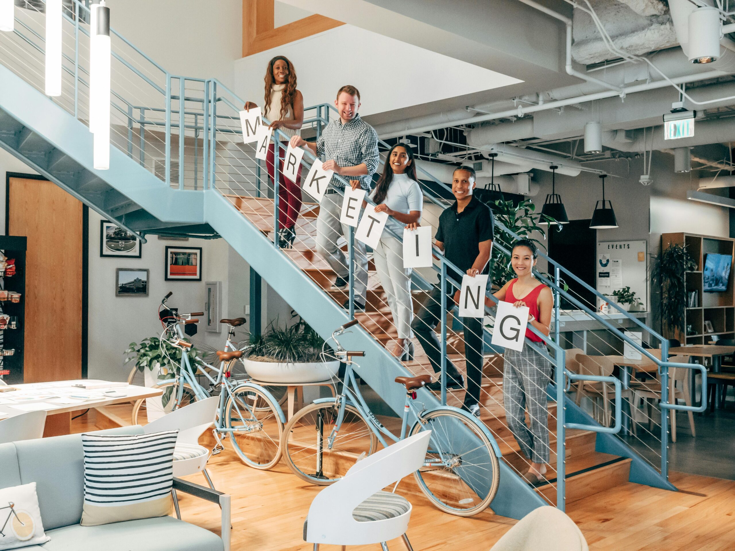 Diverse coworkers holding 'MARKETING' signs on staircase in modern office.